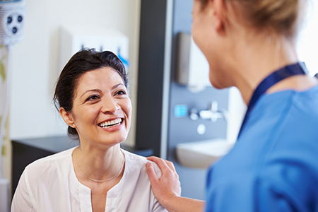 The image features two individuals in a medical setting, with a woman standing next to a man in scrubs who appears to be a healthcare professional, both smiling and engaged in conversation, suggesting a friendly interaction between them.