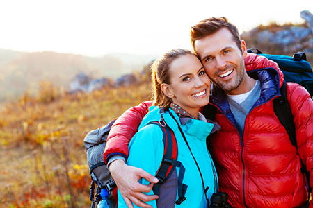 A man and woman posing together outdoors during daylight, smiling at the camera, with the man wearing a backpack and both dressed for outdoor activities.
