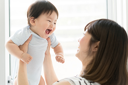 The image shows a woman holding a smiling baby close to her face, both indoors with natural light coming from behind them.